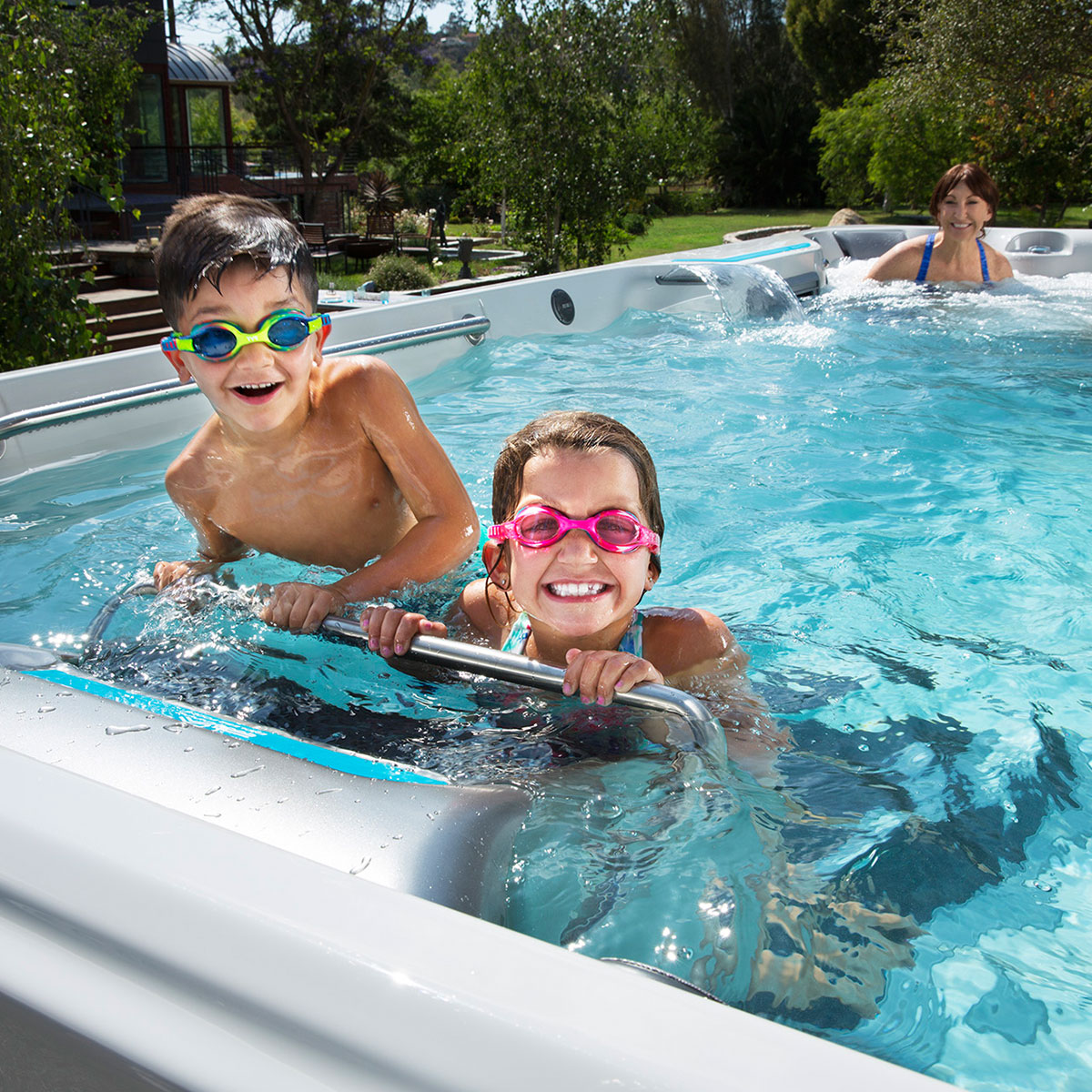 Two children with goggles smile in a backyard pool, while a person in the background relaxes in the water. Bright, sunny day.