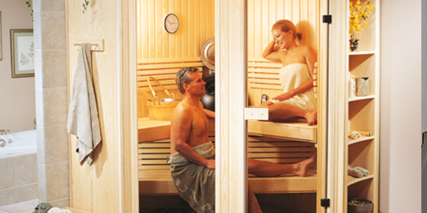 Two people relax in a wooden sauna. Towels hang nearby. Shelves hold various items, and a clock is mounted on the wall.