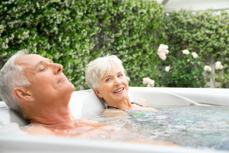Two people relaxing in an outdoor hot tub, surrounded by lush green foliage and blooming white flowers, enjoying a serene moment.