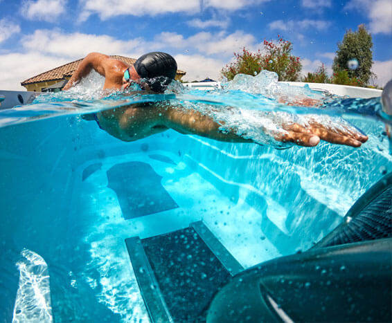 A person swimming in a clear blue pool on a sunny day, wearing goggles and a cap. A house and trees are visible.