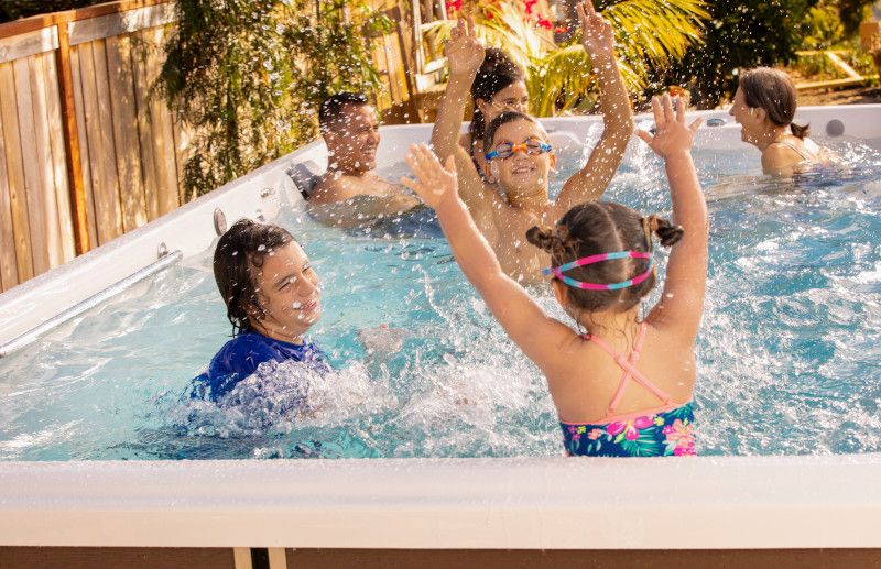 A group of people and children play joyfully in a backyard pool, splashing water and enjoying the sunny outdoors, surrounded by plants and a fence.
