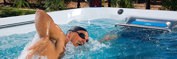 A person swims in a pool with goggles and a swim cap, beside a timer displaying 1:22, surrounded by a garden area.