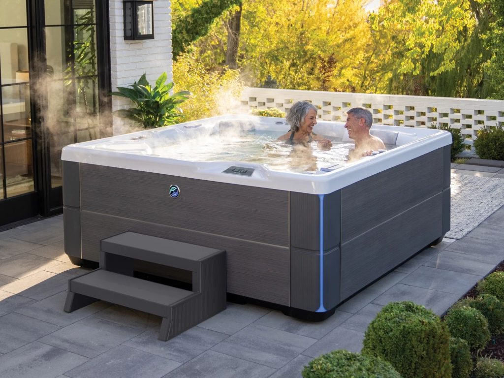 Two people are relaxing in a steaming hot tub on a patio, surrounded by lush greenery and a modern brick house exterior.