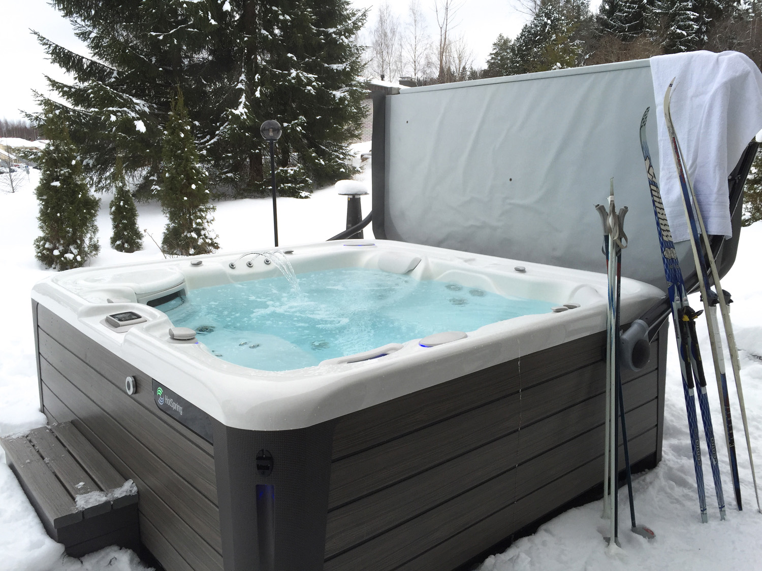 A bubbling hot tub sits outdoors in a snowy landscape, surrounded by evergreen trees, with ski equipment leaning against it.