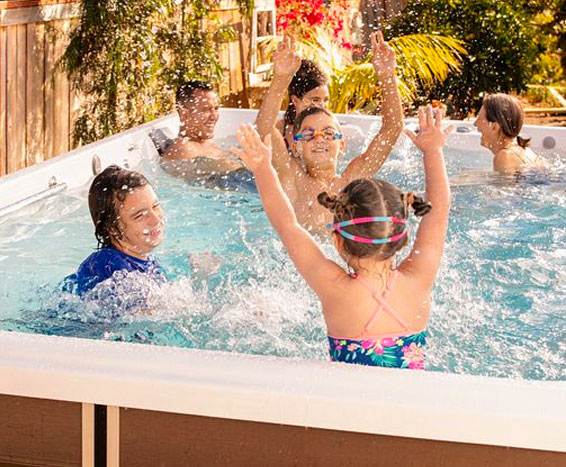 A group of people, including children, enjoys a backyard pool on a sunny day, splashing water and having fun near wooden fencing.