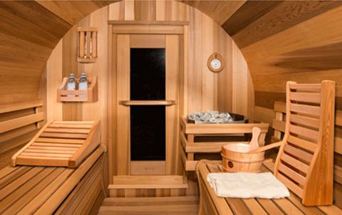 A wooden sauna interior with benches, a door, stones, a bucket, and towels. Shelves hold small bottles. Walls and ceiling are curved.