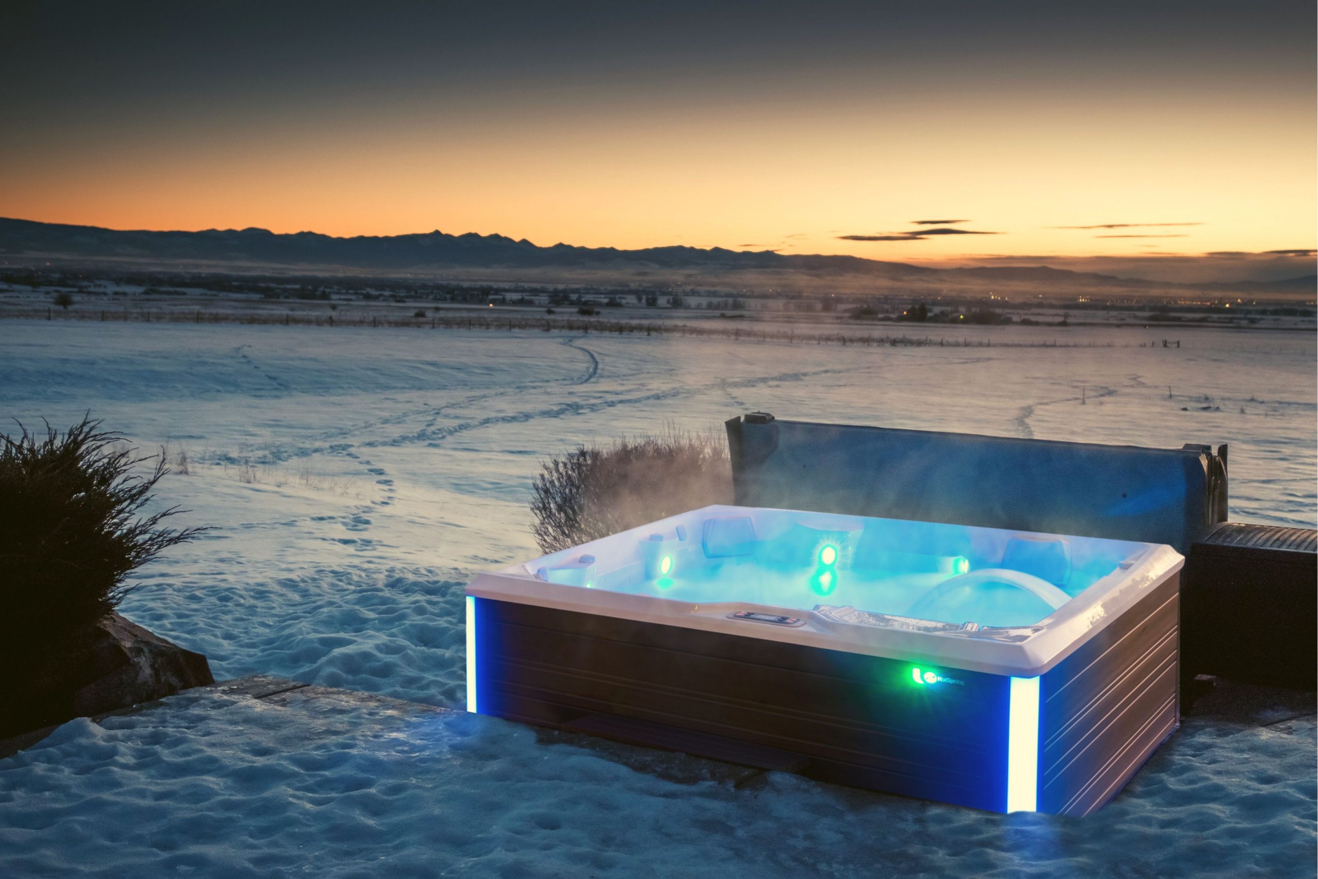 A steaming hot tub overlooks a vast, snow-covered landscape at sunset, with distant mountains providing a serene, picturesque backdrop.
