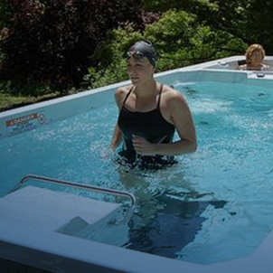 A person in swimwear and cap walks in an outdoor pool, surrounded by lush greenery and trees on a sunny day.