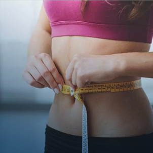 A person wearing a pink top measures their waist with a tape measure, focusing on fitness or body measurements in a well-lit room.