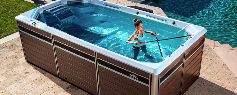 A person exercises in an outdoor swim spa with resistance bands. The spa is framed by stone tiles and surrounded by a lawn.