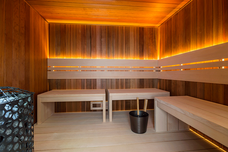 A wooden sauna interior with glowing ambient lighting, wooden benches, a heater, and a bucket with a ladle on the floor.