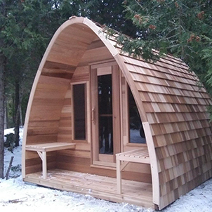 A small, wooden, arched cabin with shingled roof sits in a snowy forest. It features a door, window, and two benches.