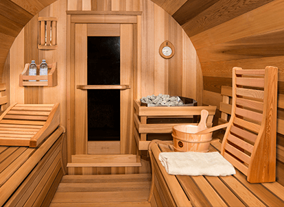 Wooden sauna interior with benches, water bucket, towel, clock, and stones. Warm lighting highlights the cozy atmosphere. No people or landmarks present.