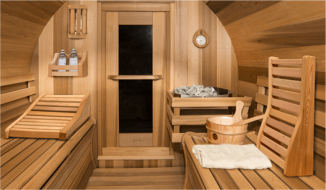 A wooden sauna interior with benches, a bucket, ladle, and stones. Soft lighting creates a relaxing atmosphere with towels and bottled water.