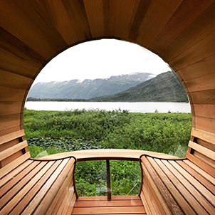 A round wooden sauna with a lake and mountain view. The interior features benches, offering a relaxing nature scene through a circular window.